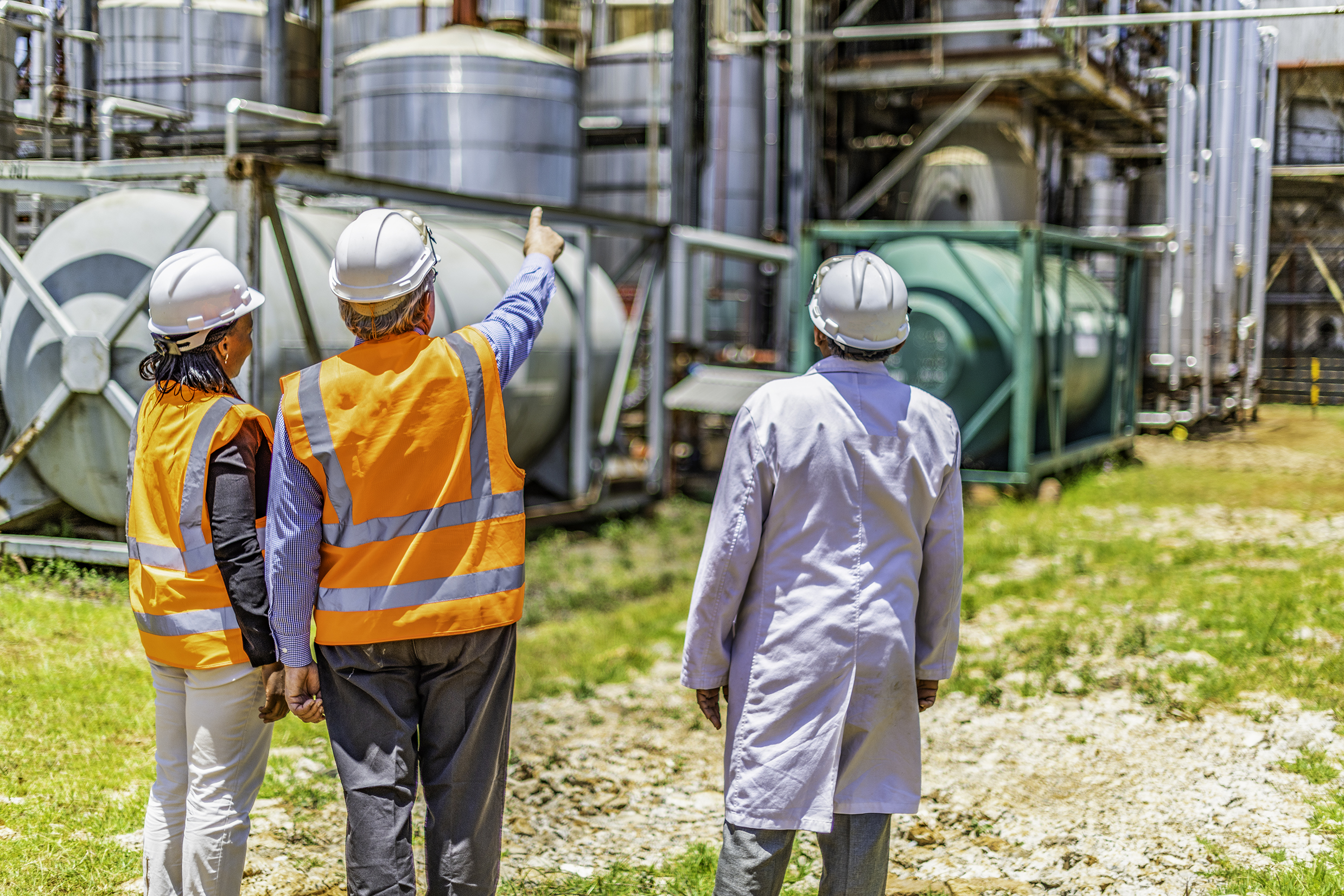 Back view of successful older business owner or manager in discussion with employees on site at a chemical plant. - Diversity of ethnic background, Caucasian, African American and Indian. age and gender. All wearing appropriate PPE, Personal Protective Equipment. Non urban location. Outdoors.