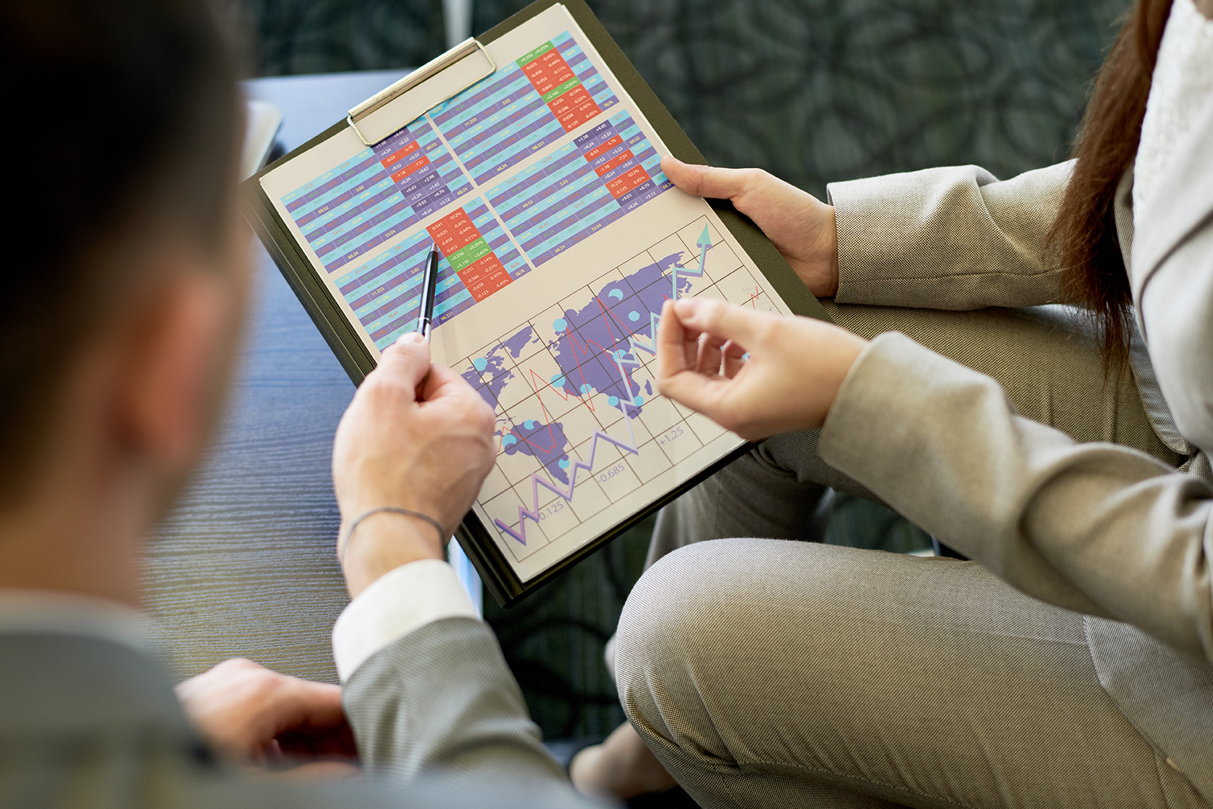 Worldwide Business Statistics - High angle close up of two business people holding graph with world map while discussing statistics in meeting and planning project,